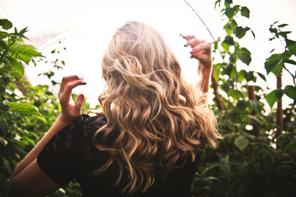 A blonde woman among greenery with her back turned to the camera.