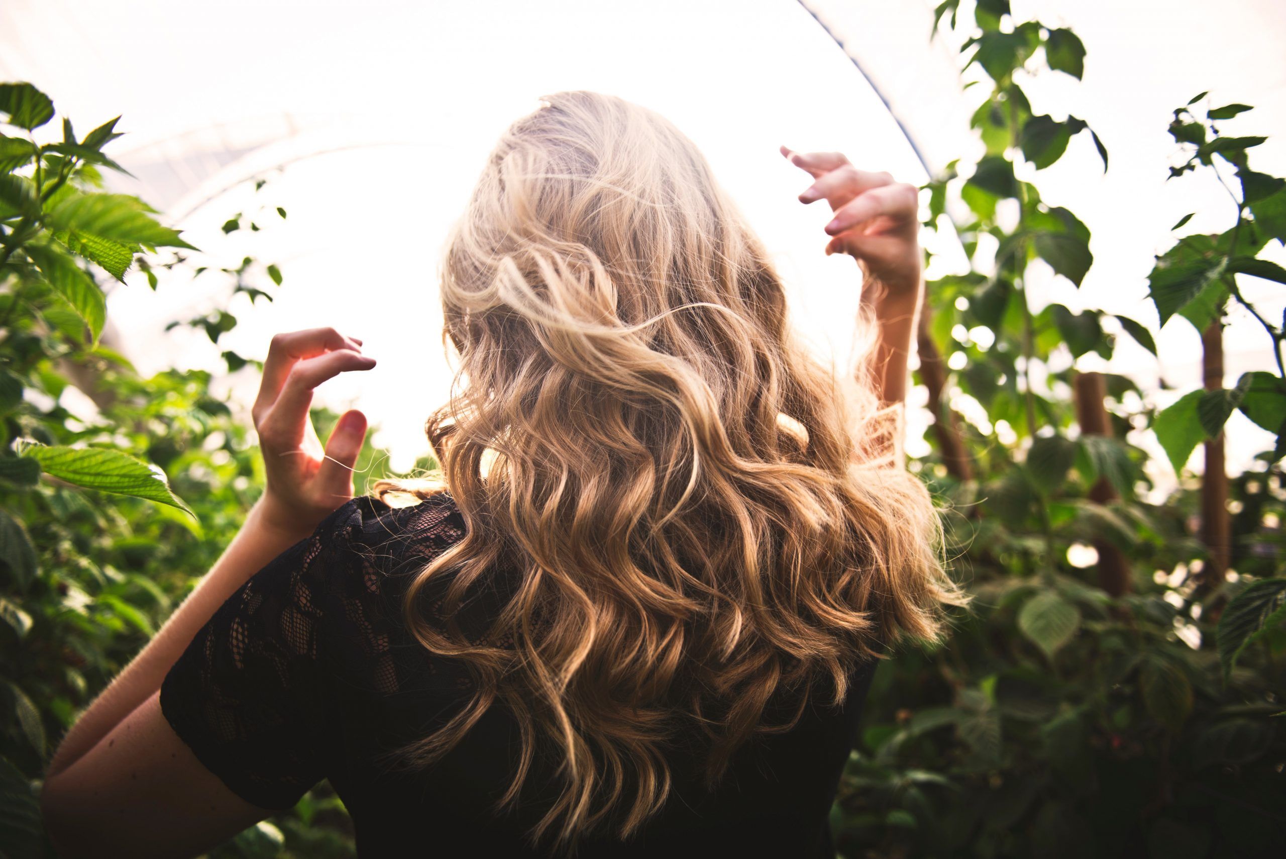 A blonde woman among greenery with her back turned to the camera.