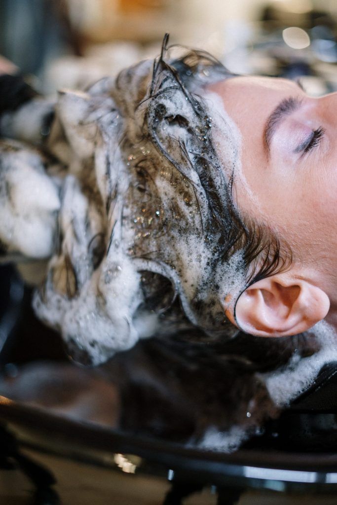 A close-up of a person receiving a shampoo wash at a salon, with thick foam lather covering their hair and neck, and their eyes closed in relaxation.