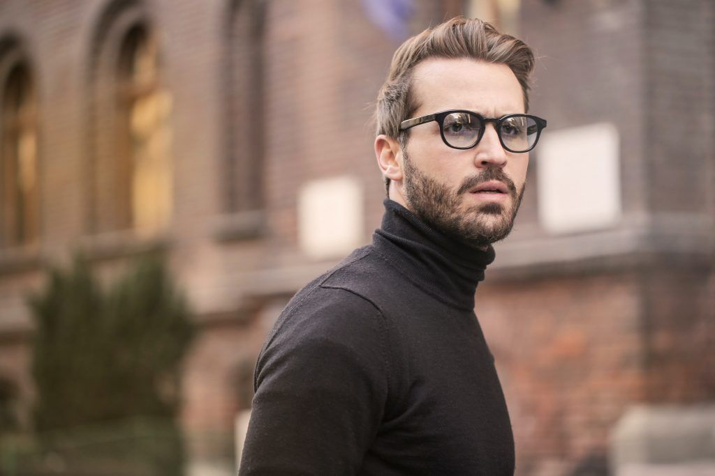 A man wearing black turtleneck jumper and black framed glasses, with brunette hair and beard looking puzzled something outside of the frame.