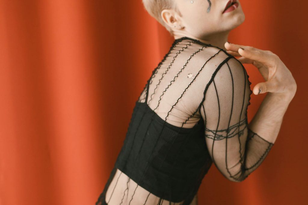An artistic close-up shot of a person wearing a sheer black top with delicate crisscross patterns. The image captures the back and shoulder, with a soft, dramatic pose, against a vibrant red background. The individual has a subtle makeup look, with a small tear-shaped design near the eye.